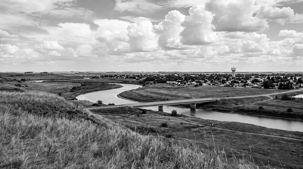 Generated black-and-white scene: a prairie river valley with a low bridge and a small city on the far bank; not a photograph of North Battleford.